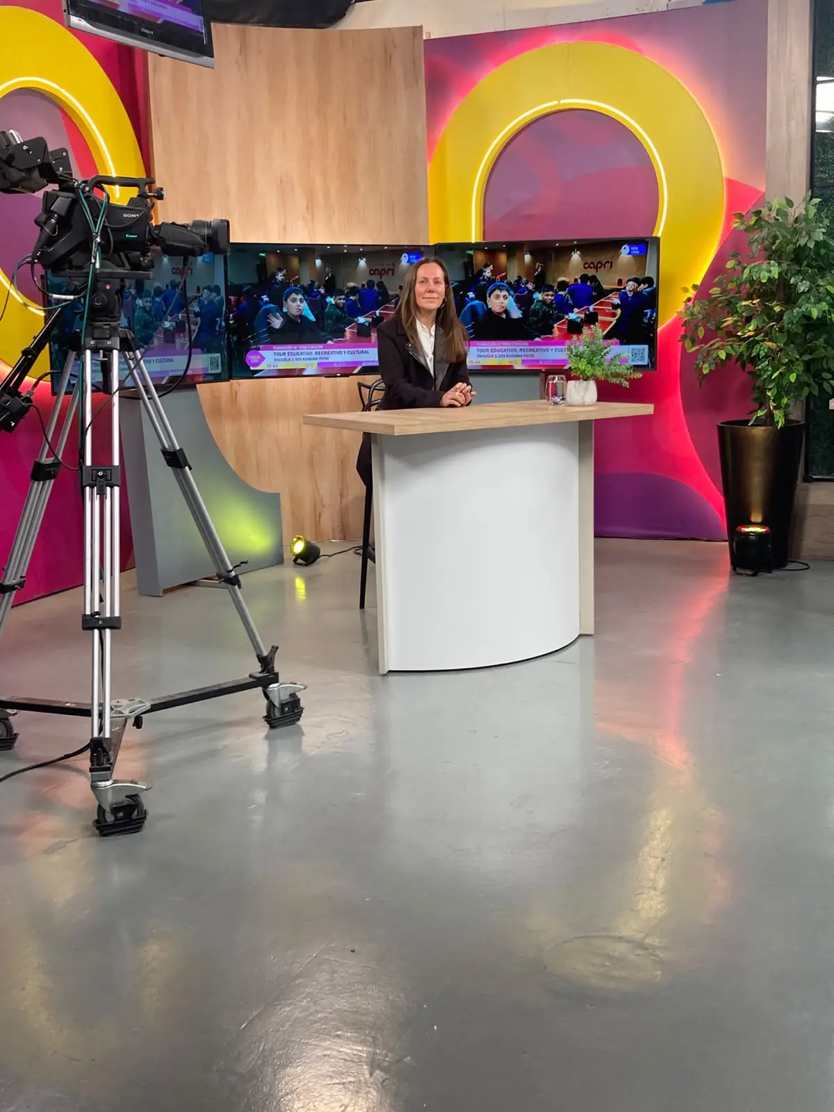 La rectora sentada a la mesa del estudio, de frente a una cámara sobre trípode; detrás, las pantallas del programa muestran a un grupo de estudiantes