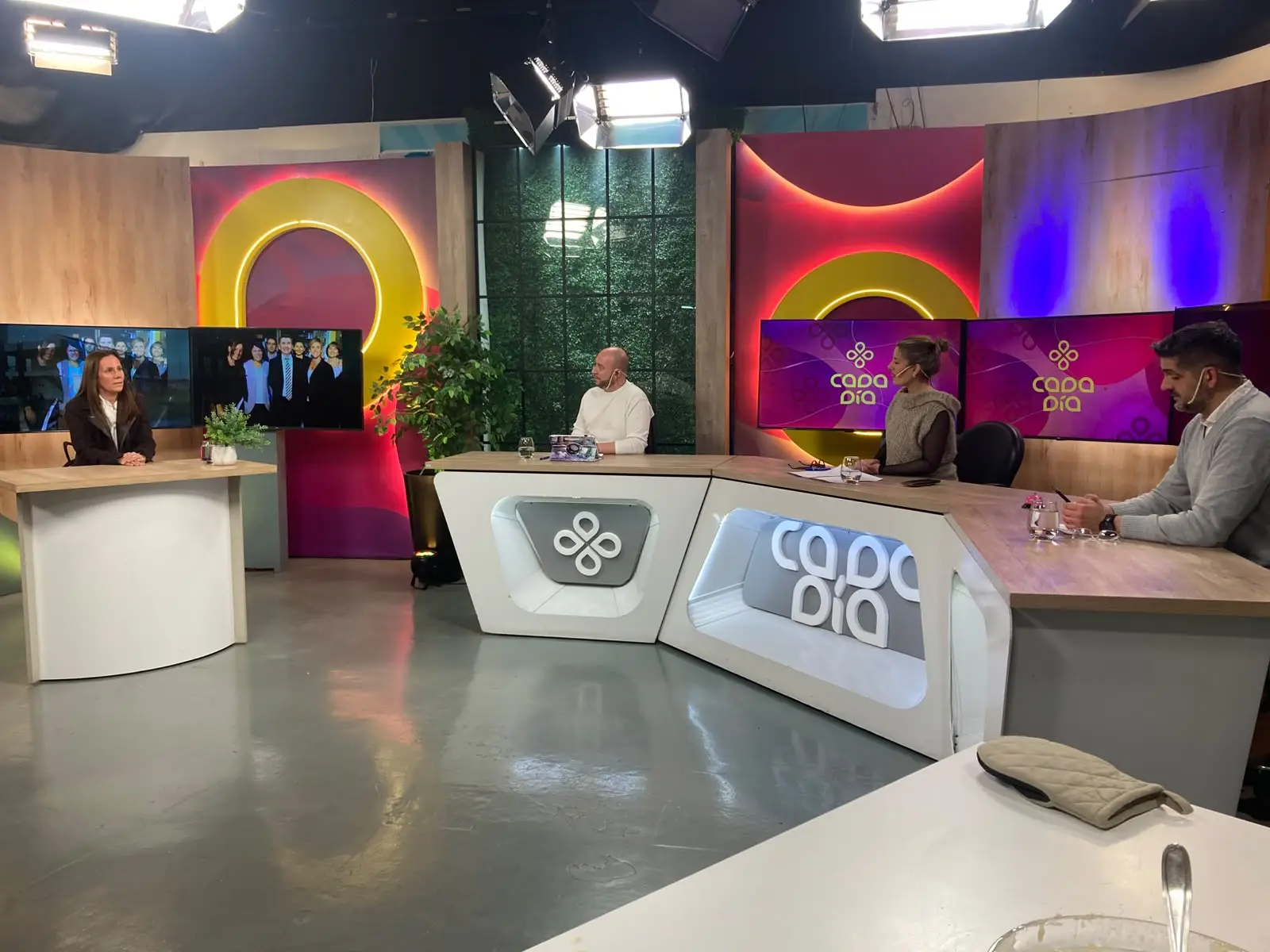 Piso del programa Cada Día: la rectora María Virginia Hernández, sentada a una mesa alta a la izquierda del estudio, escucha a los tres conductores ubicados detrás del escritorio con el logo del programa